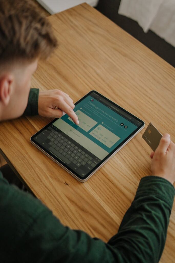 An adult male using a tablet and credit card for online shopping at a wooden desk indoors.